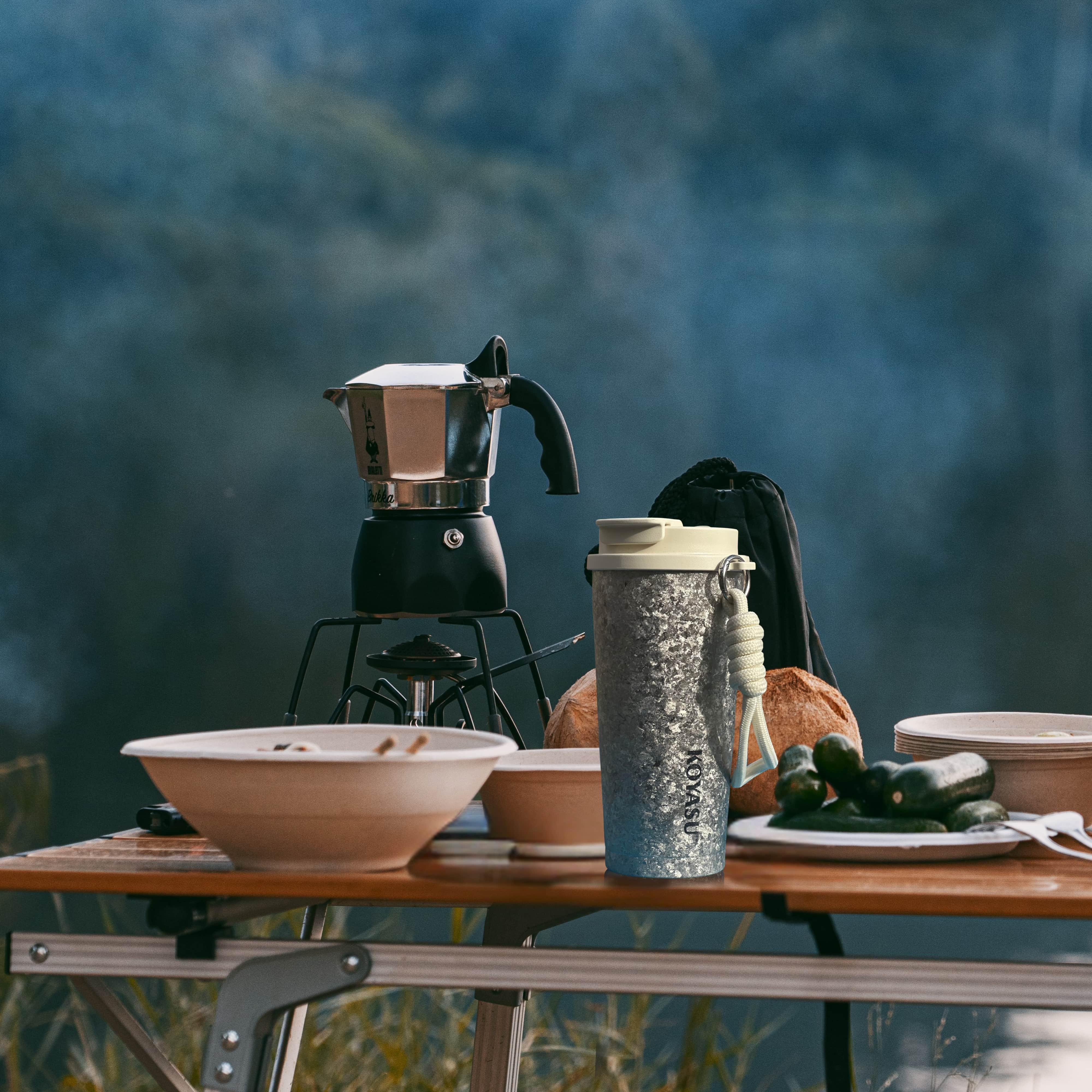 KOYASU 16oz whole-body titanium travel mug on a campsite breakfast table beside a moka pot, bowls, and fresh food. Set against a misty lake backdrop, the image emphasizes the mug’s suitability for outdoor use — lightweight, durable, and ideal for hot drinks in nature.