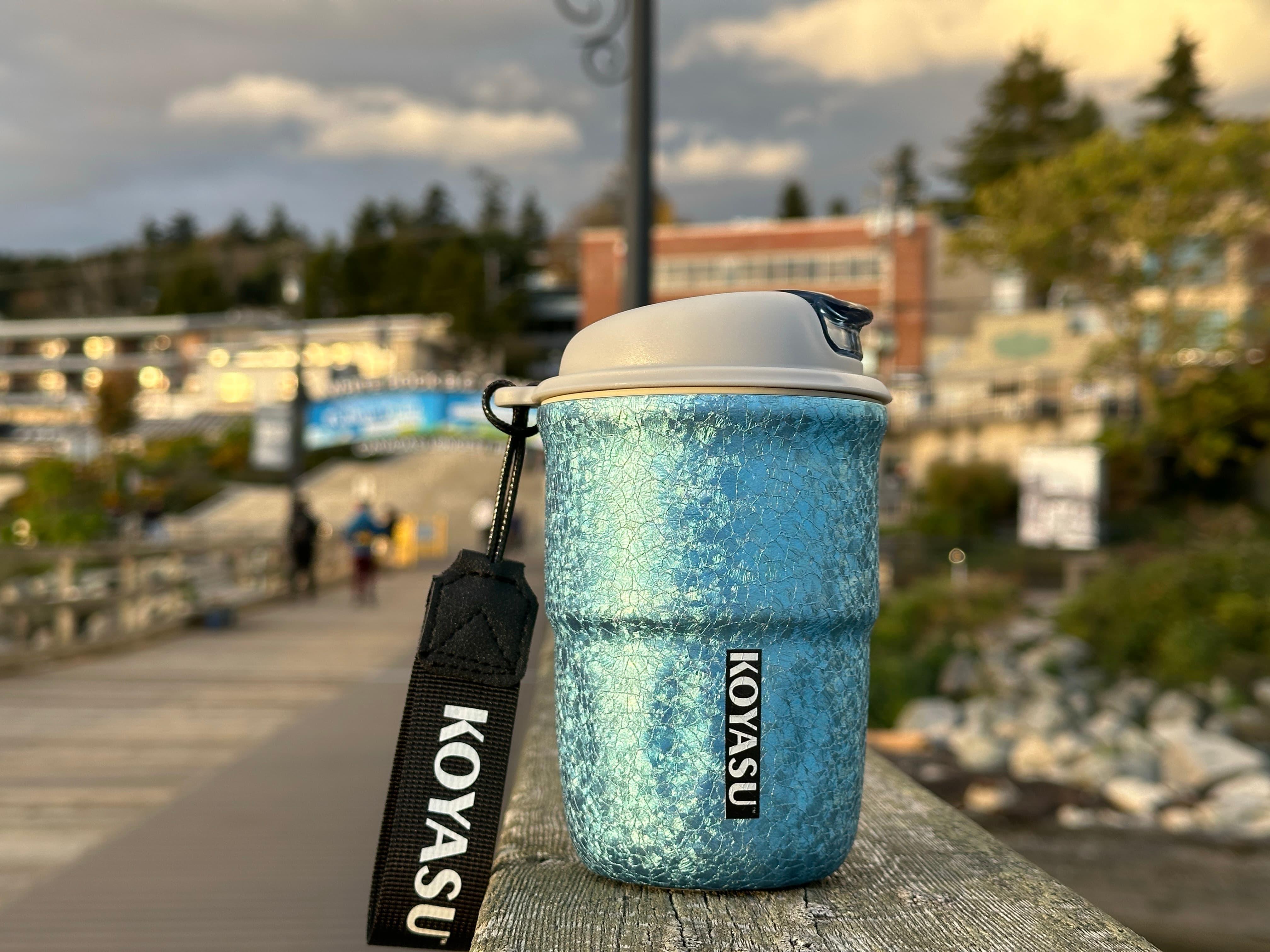 KOYASU 12oz titanium-lined mug in cool blue resting on a boardwalk railing during sunset, ideal for outdoor strolls and scenic moments by the water.