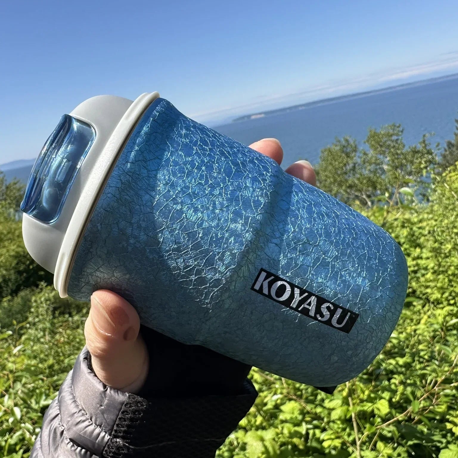 KOYASU 12oz titanium-lined mug in Cool Blue, shown in outdoor coastal setting, hand-held against the ocean backdrop