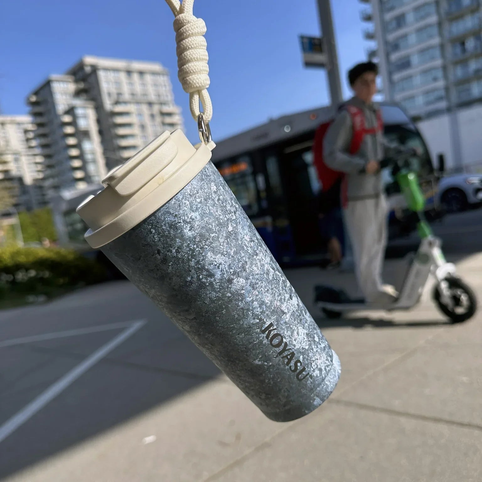 KOYASU 16oz Whole-Body Titanium Mug hanging by rope strap in an urban commuting scene with bus and scooter