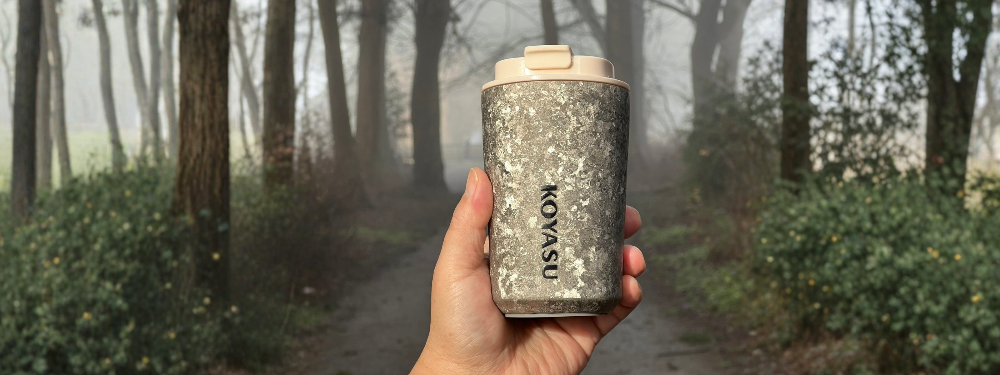 KOYASU® Ti-Zen™ 12oz pure titanium coffee mug held by hand on a misty forest trail. Ultralight, double-wall vacuum insulated travel tumbler for camping and daily commute, featuring raw crystal titanium texture and leakproof lid.