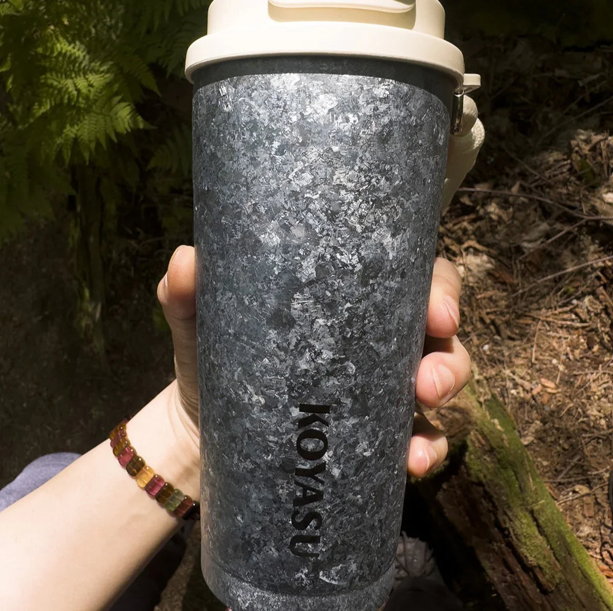 KOYASU 16oz titanium mug in outdoor forest setting, hand-held with visible raw titanium texture — lightweight and ideal for hiking