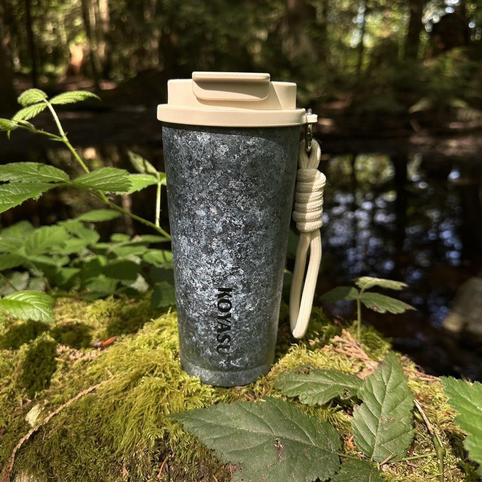 KOYASU pure titanium travel mug resting on a wet mossy rock beside a forest stream. Ultralight, double-wall insulated camping coffee tumbler for outdoor hiking and survival gear.